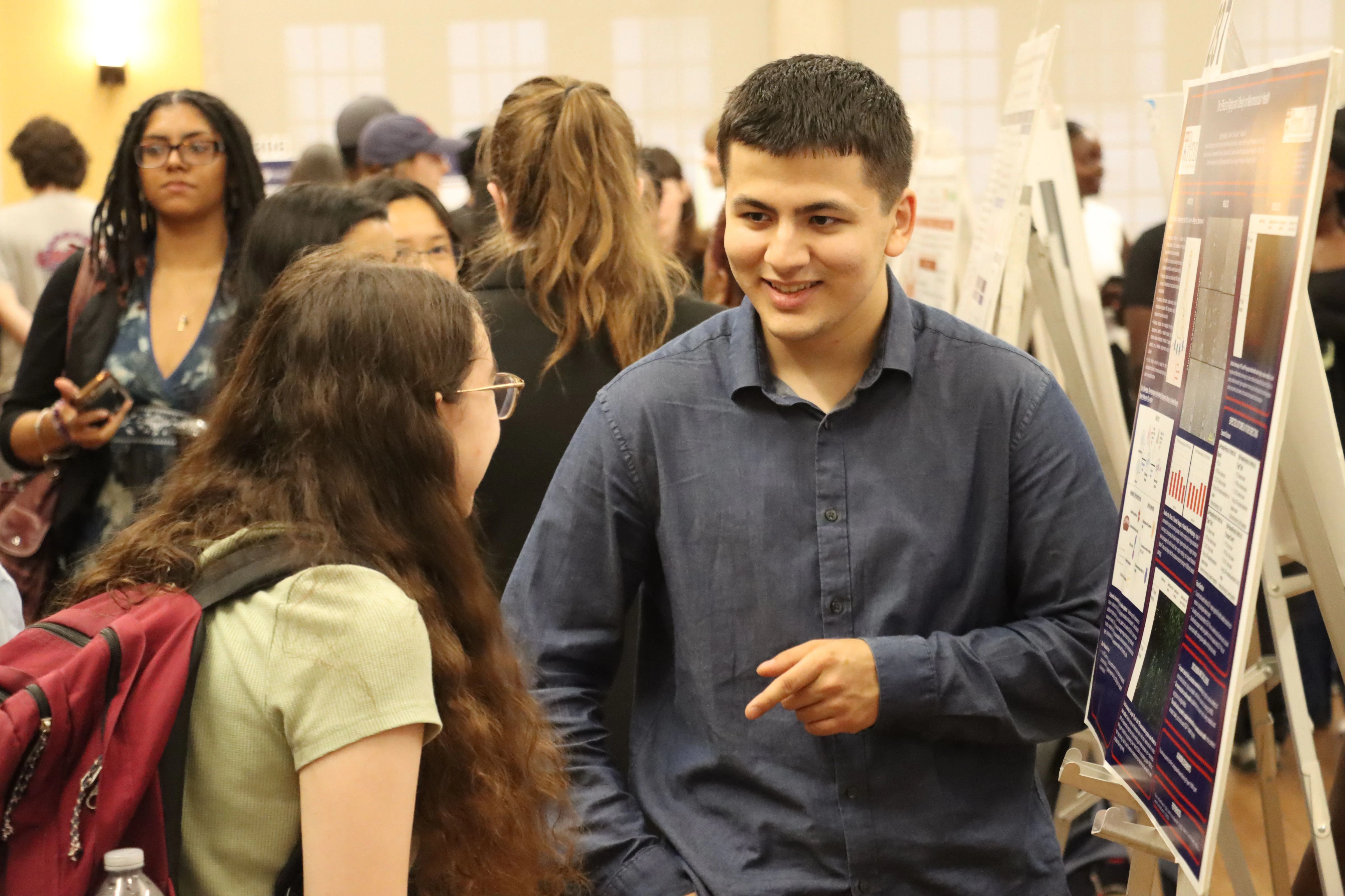male student standing at a presentation board, sharing with another student about his project